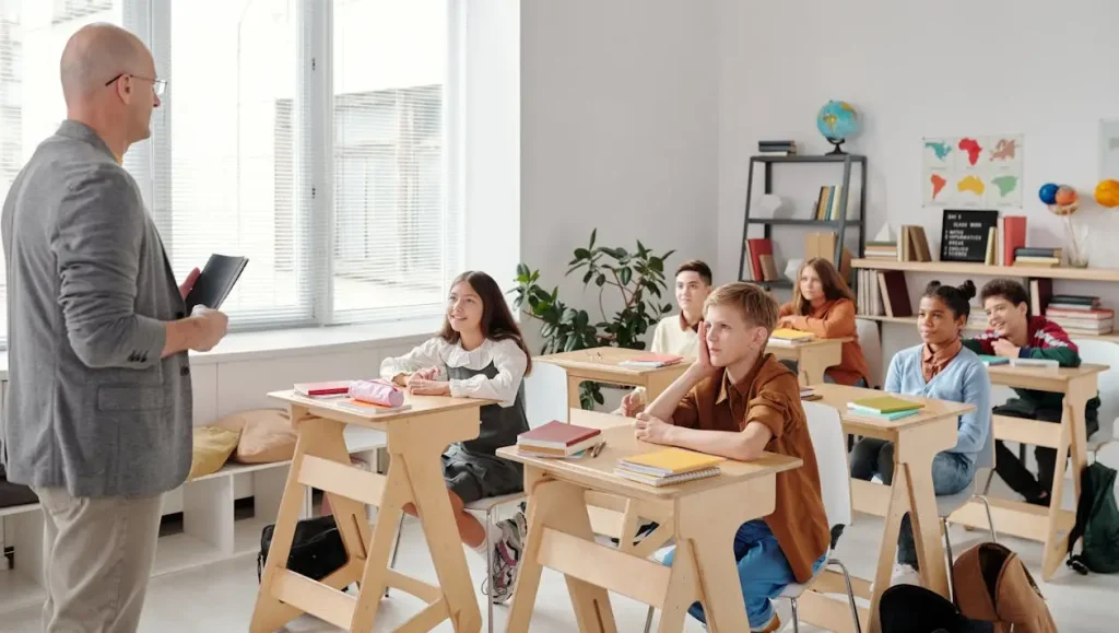 Teacher standing in front of attentive students seated at wooden desks in a bright classroom