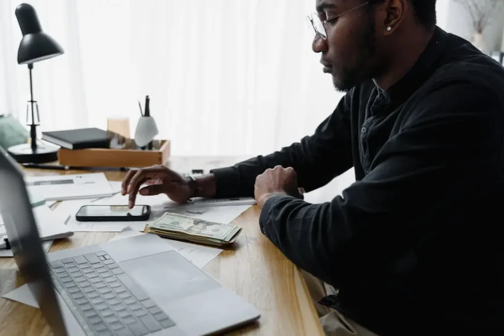 Man using smartphone at wooden desk with laptop and money in bright office