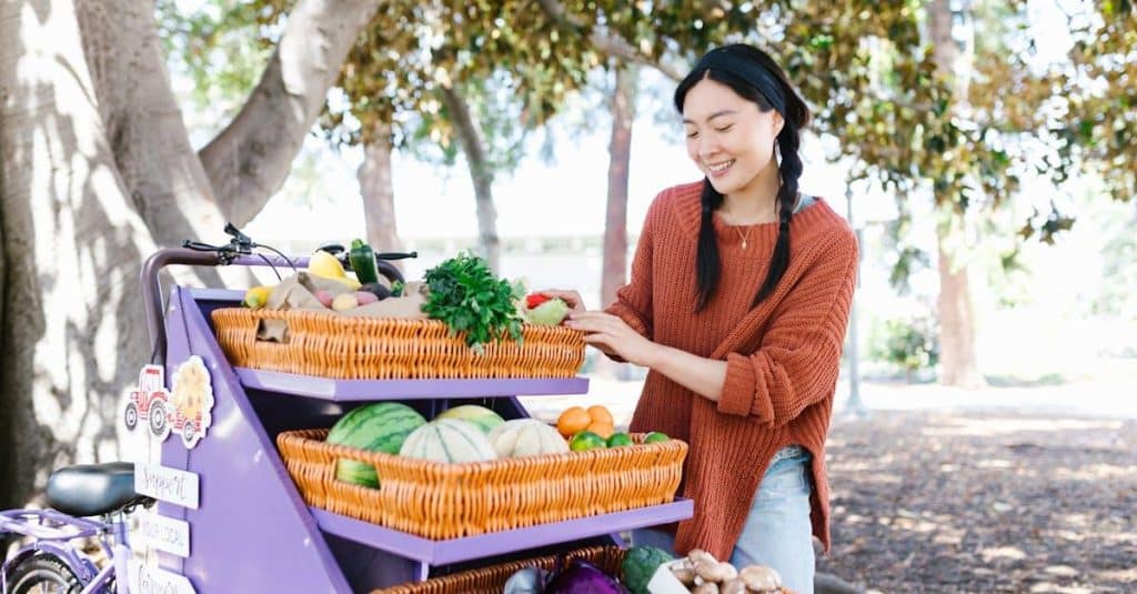 Woman in sweater selecting fresh produce at outdoor market fruit stand.