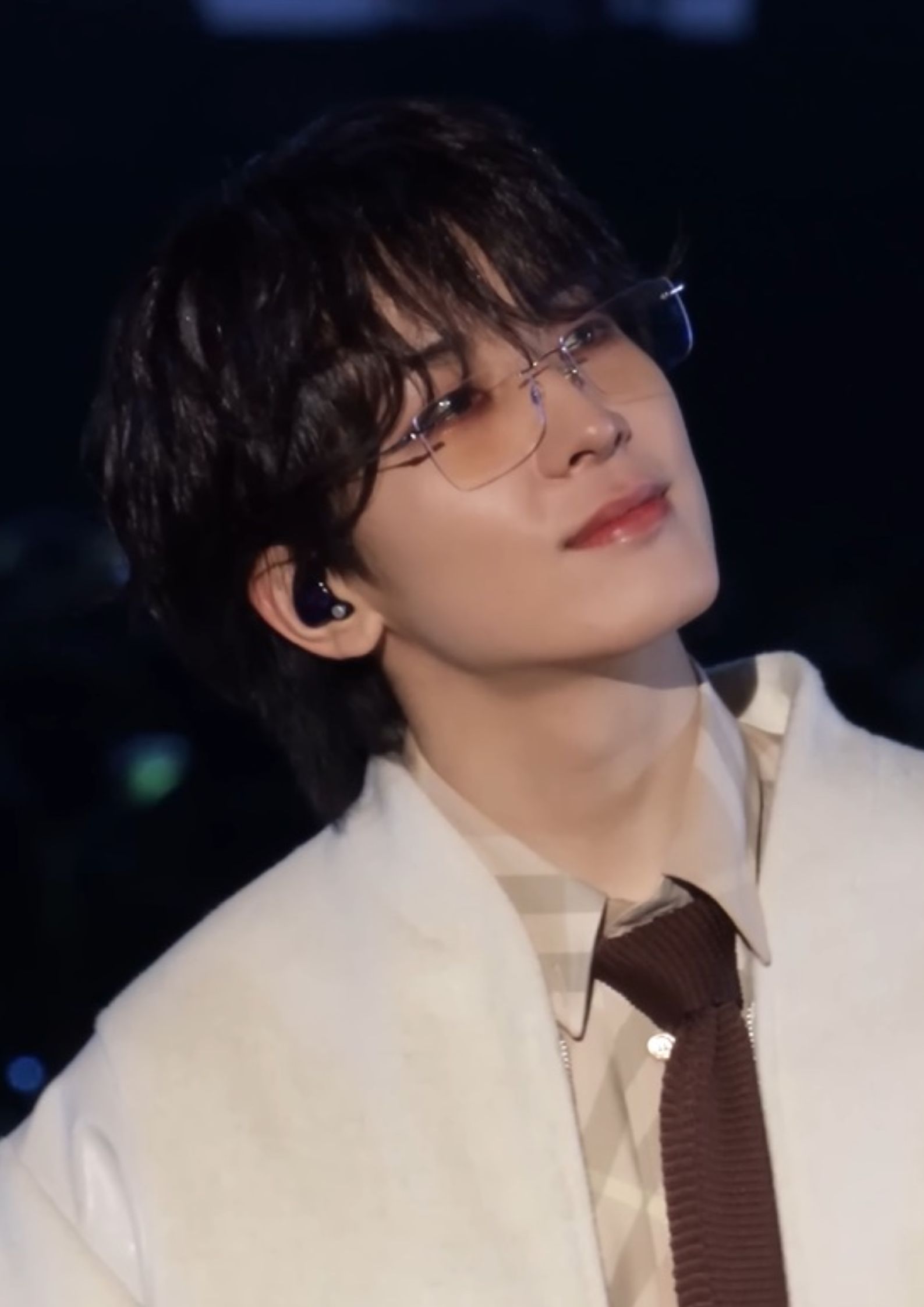 Wonwoo of Seventeen performing on stage wearing glasses, a beige shirt, brown tie, and white jacket, looking thoughtful.