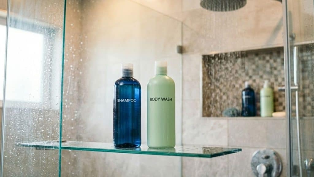 An elegant, modern glass shower stall featuring a blue bottle labeled SHAMPOO and a light green bottle labeled BODY WASH sitting side by side
