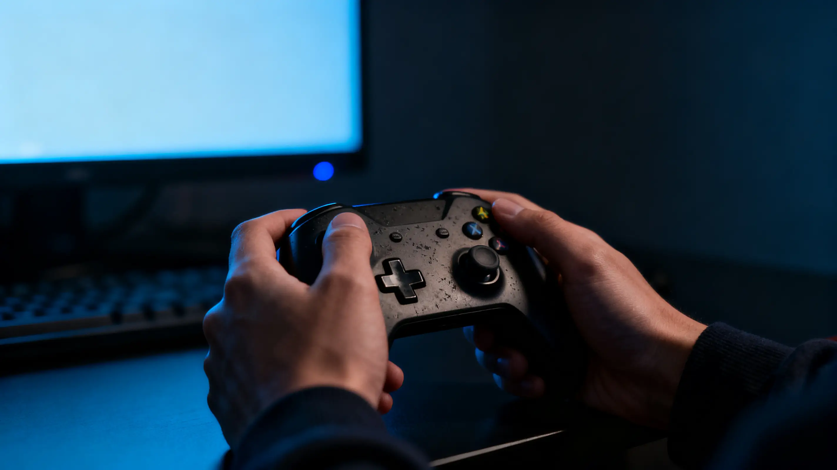 Hands holding a black gaming controller in front of a computer screen in dim lighting