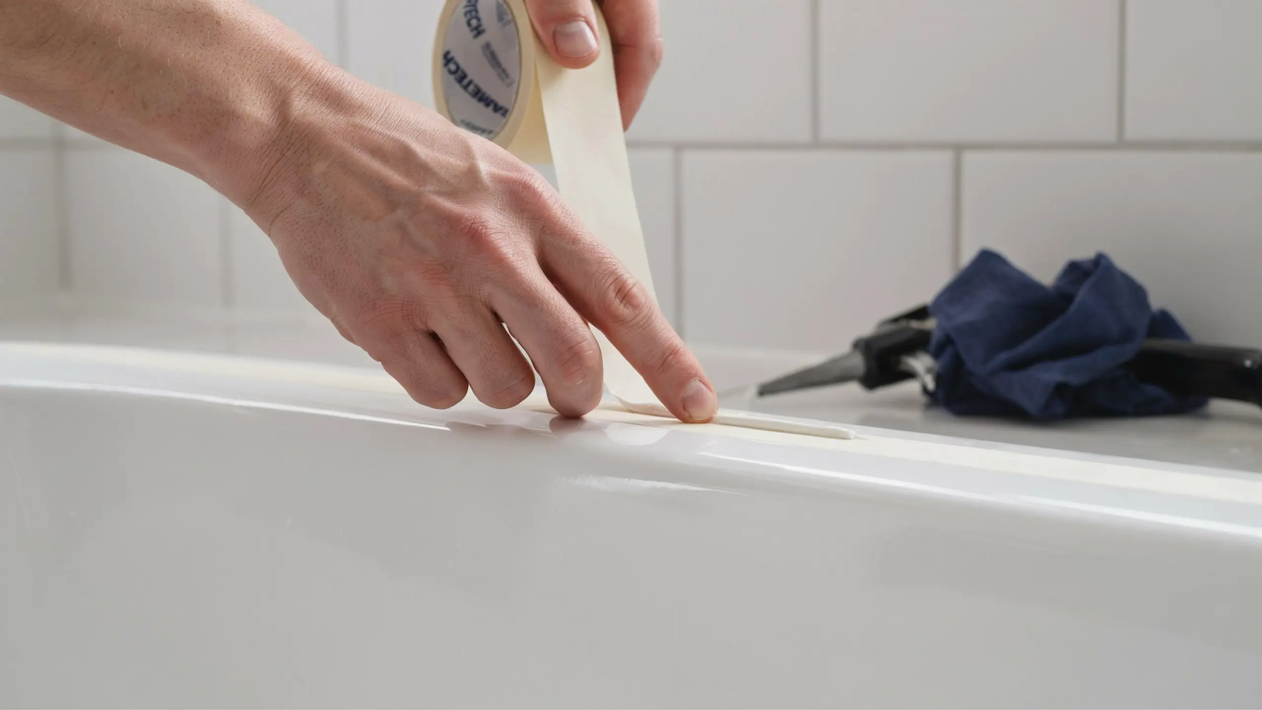 Hands applying painter’s tape along bathtub edge to create straight caulk line.