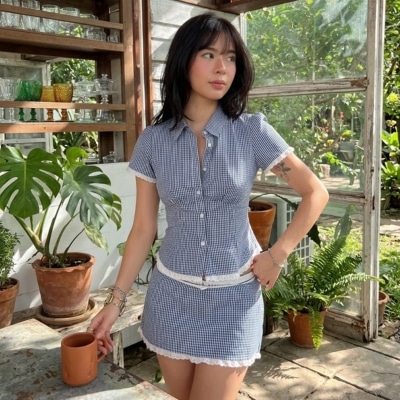 Woman wearing a blue checkered matching top and skirt holding a mug in a sunlit greenhouse.