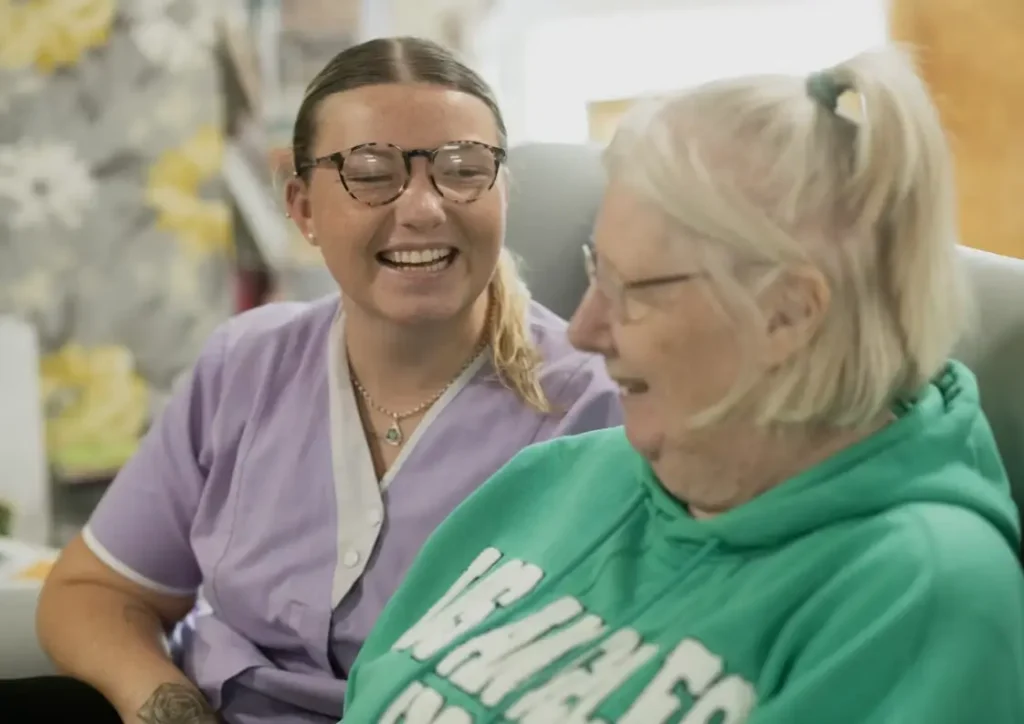Caregiver smiling with elderly woman in nursing home, both seated and engaged in conversation