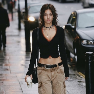 Woman in black cropped top and cargo pants walks on wet city street holding a coffee cup.