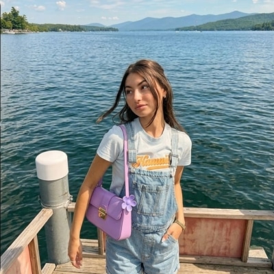 Young woman in denim overalls standing on a dock by a calm lake under a blue sky.