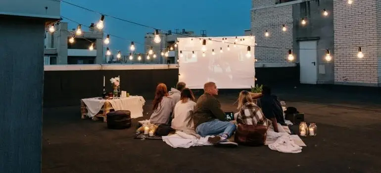 Group of people watching outdoor movie on rooftop with string lights and blankets at dusk