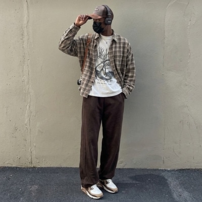Person wearing a plaid flannel shirt, graphic tee, and dark pants standing against a beige wall.