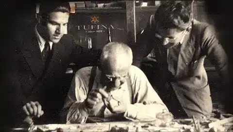Elderly watchmaker examining components with magnifying glass surrounded by two young men in workshop