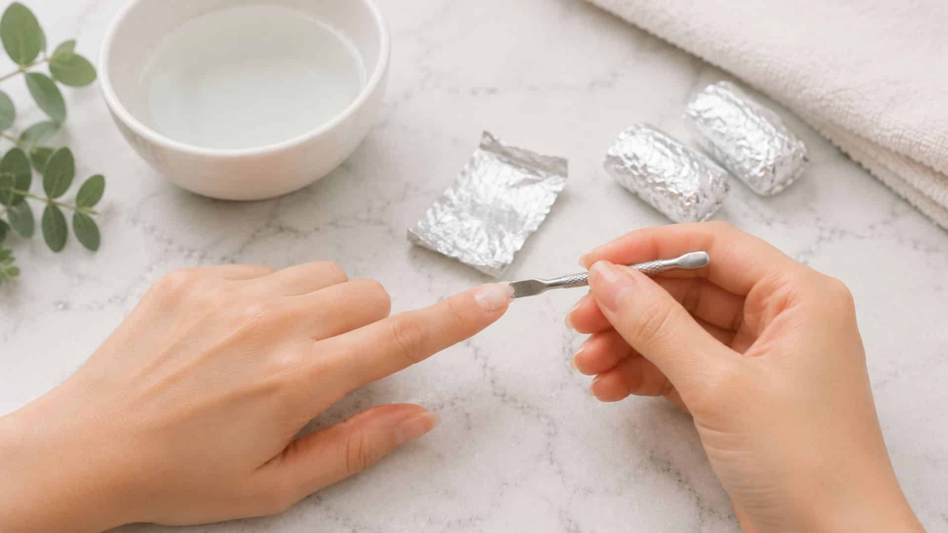 person gently lifting softened acrylic with a cuticle pusher beside foil wraps on a marble surface