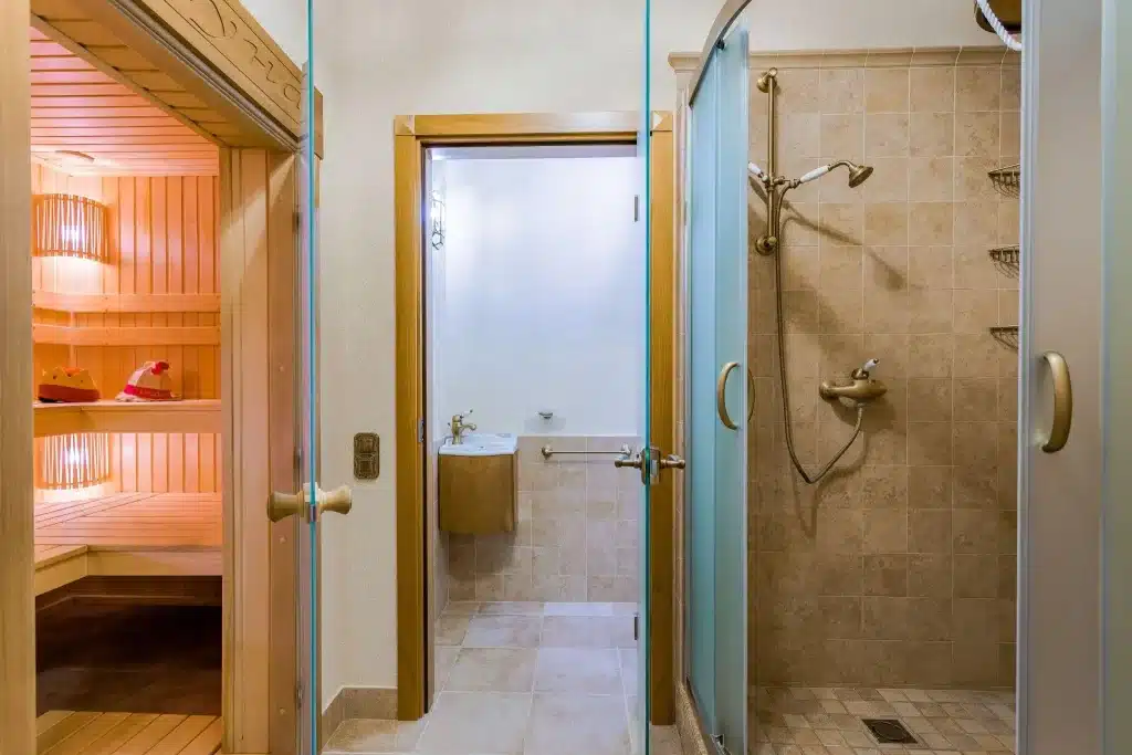 Sauna room with wooden benches beside a tiled shower and small sink in a modern bathroom