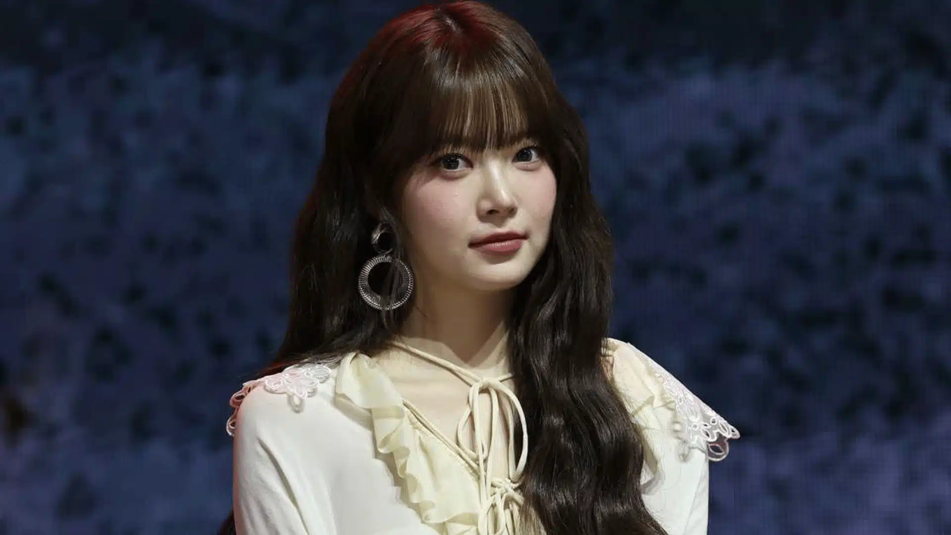 Female K-pop idol on stage in cream lace outfit with long dark hair and bangs, wearing circular earrings under blue concert lighting.