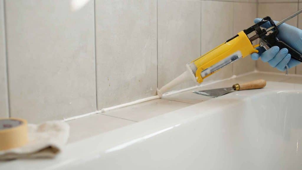 Hand applying silicone caulk along bathtub edge where tile meets tub, sealing joint with caulking gun