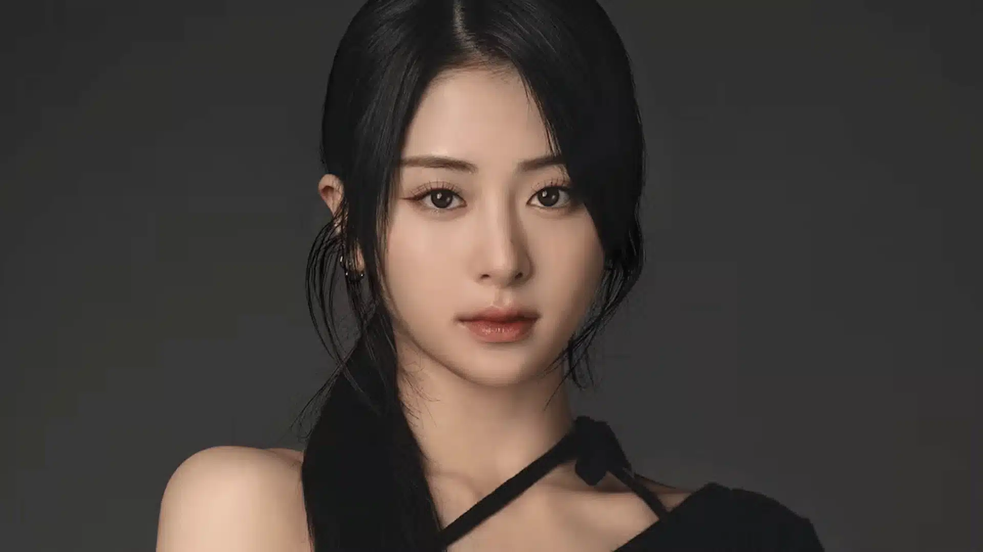 Female K-pop idol in close-up portrait against a dark grey background, wearing a black asymmetrical outfit, with a calm direct gaze and low ponytail hairstyle.