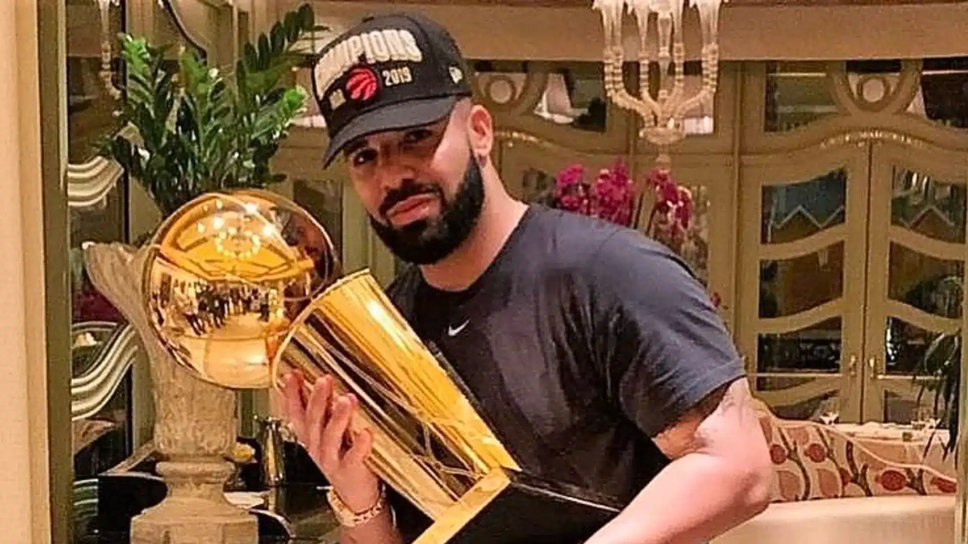 Drake holding a championship trophy and commemorative basketball in an elegant indoor setting, wearing a celebratory champions