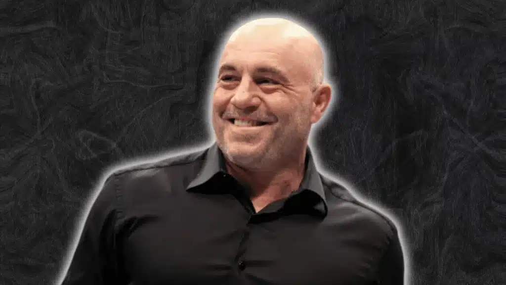 Joe Rogan in a black shirt smiling while looking slightly to the side against a dark textured background.