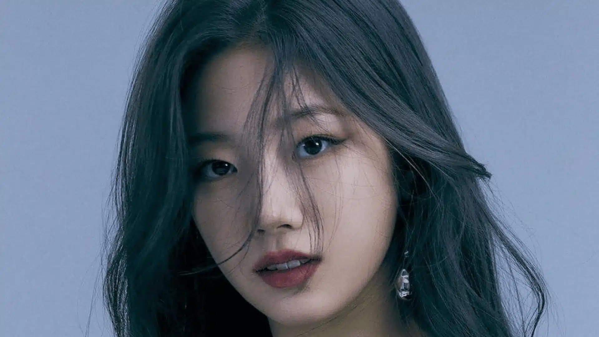 Female K-pop idol in close-up portrait with soft blue-toned lighting, long dark hair partially covering face, wearing earrings and a calm expression.