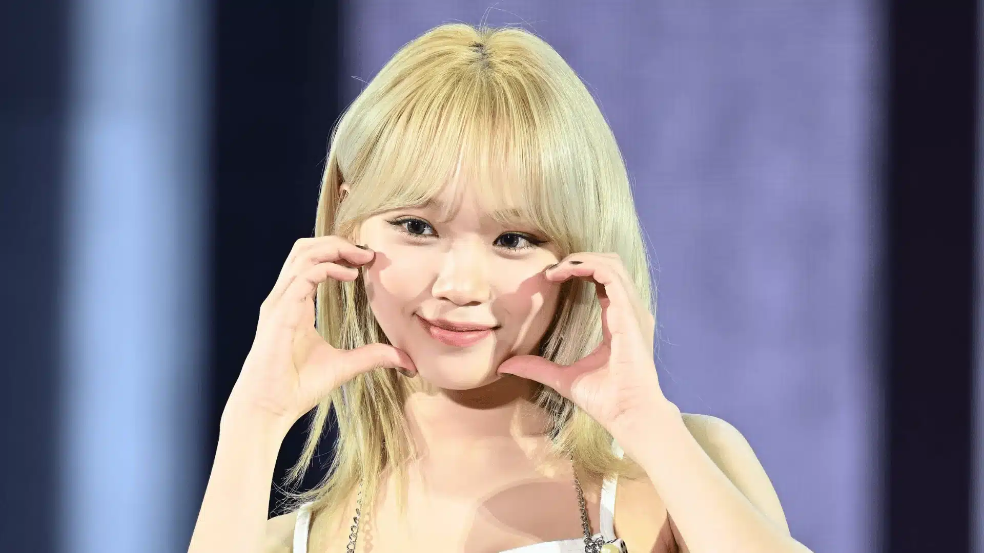 Blonde K-pop idol in close-up on stage, smiling and forming a heart shape with both hands under bright performance lighting.
