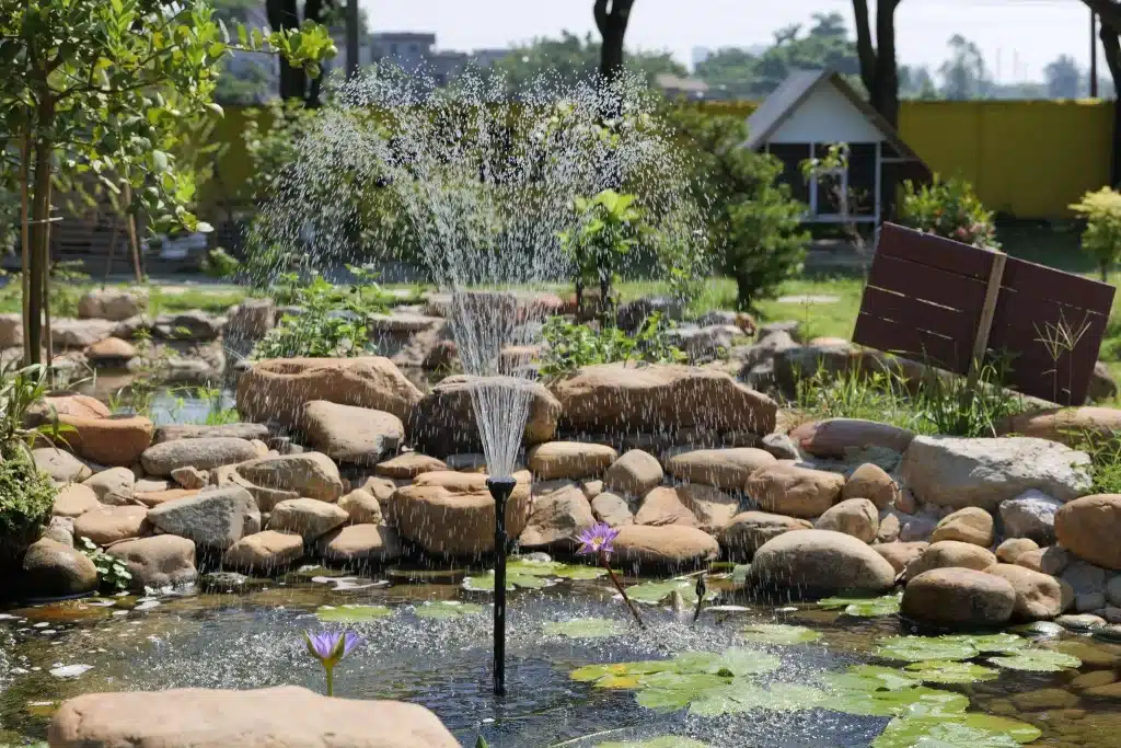 Garden pond with fountain and water lilies surrounded by rocks under sunny sky
