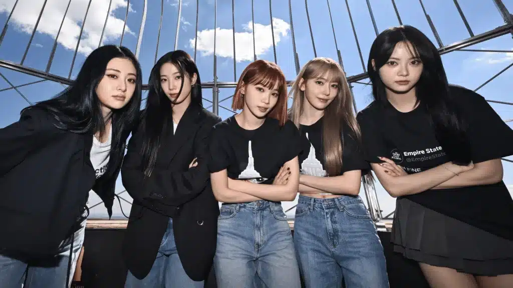 Five-member K-pop girl group posing outdoors behind metal framework, arms crossed, wearing black outfits under blue sky.