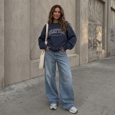 Woman wearing a navy sweatshirt and wide-leg jeans standing on a city sidewalk with white sneakers and a tote bag.