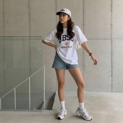 Woman wearing a white graphic T-shirt, denim shorts, white sneakers, and a baseball cap standing indoors near a concrete wall.