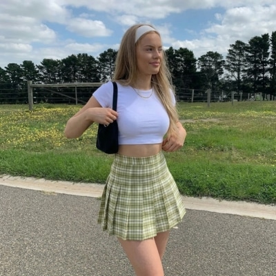 Young woman wearing a green plaid skirt and white crop top standing by a grassy field.