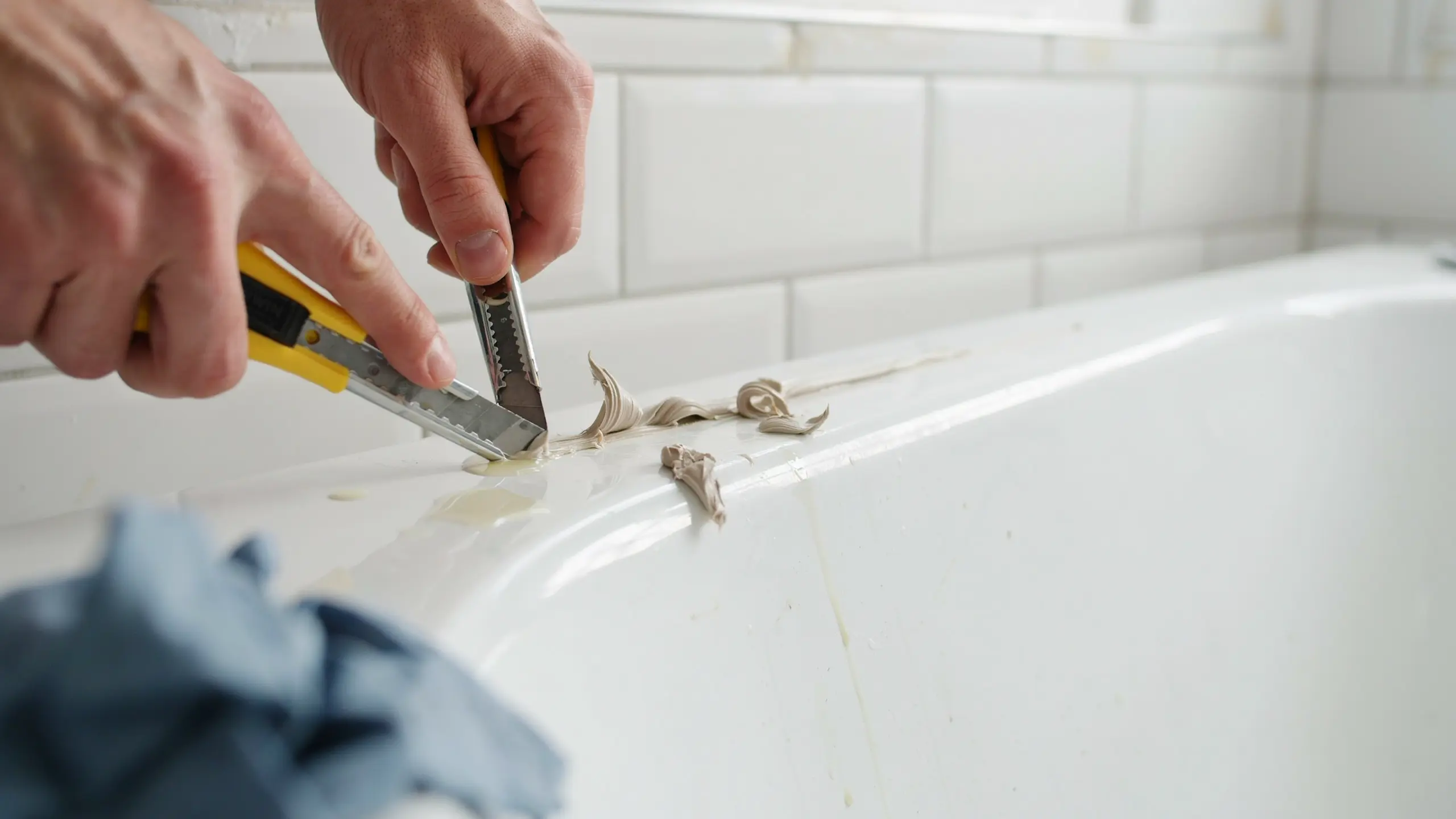 Hands scraping old caulk from bathtub edge with utility knife and removal tool.