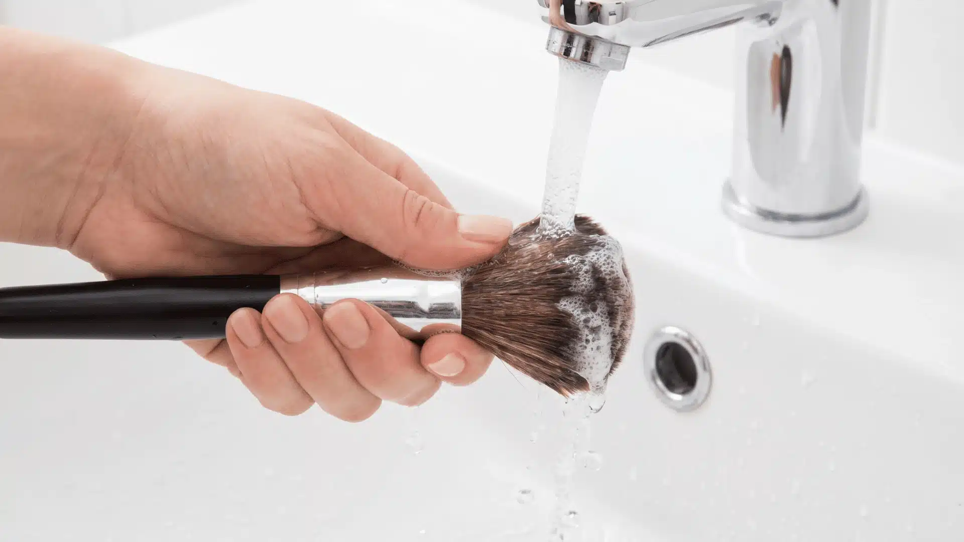 A makeup brush being rinsed under lukewarm water while excess liquid is gently squeezed from the bristles.
