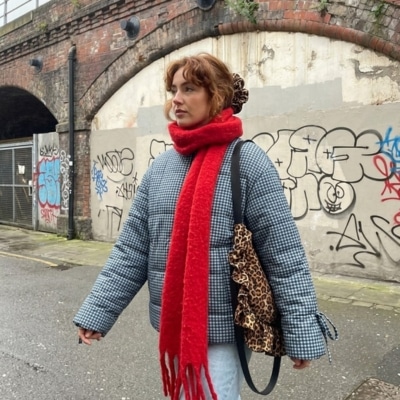 Woman wearing a checkered coat, bright red scarf, and carrying a leopard-print bag walking in an urban alley with graffiti and brick walls.
