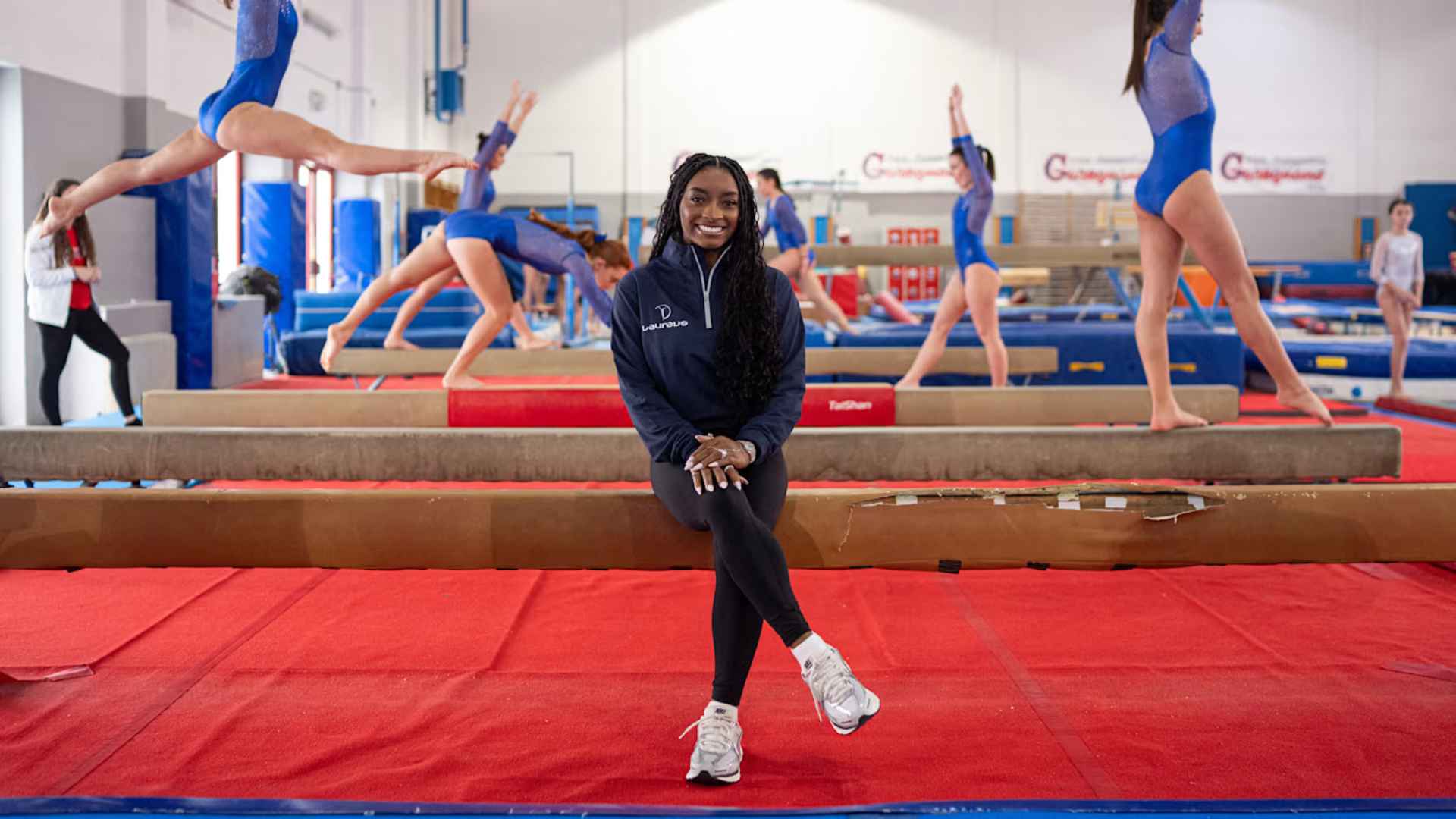 Simone Biles smiling in gym with gymnasts practicing behind