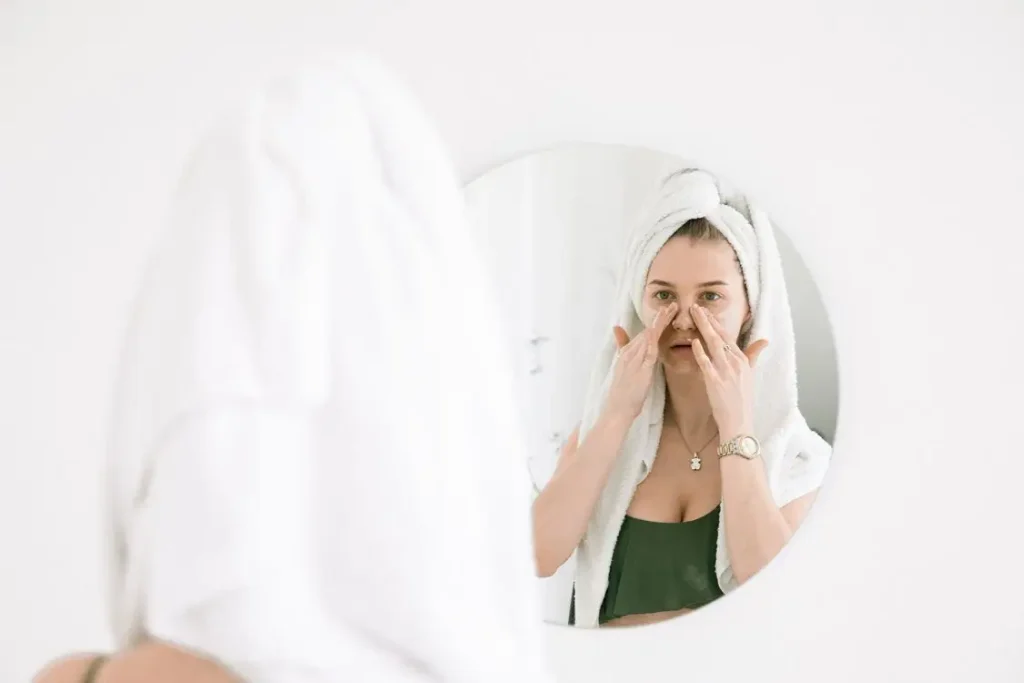 Woman with towel on head applying skincare cream in front of bathroom mirror