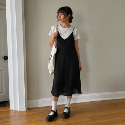 Woman in black slip dress over white T-shirt posing indoors on wooden floor with minimalist accessories