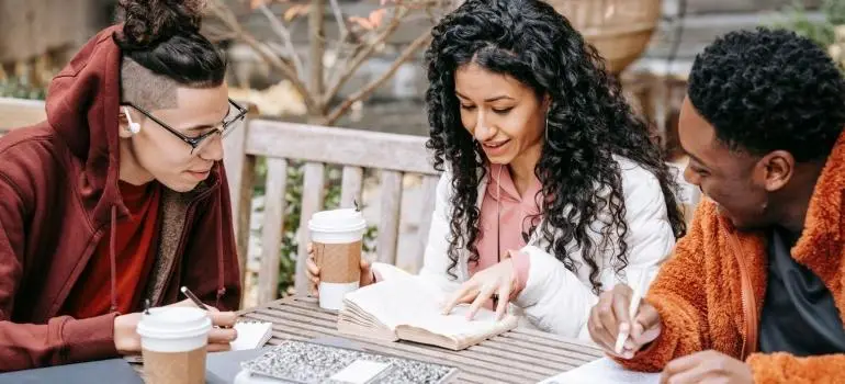 Three friends studying at a wooden table outdoors with notebooks and coffee cups