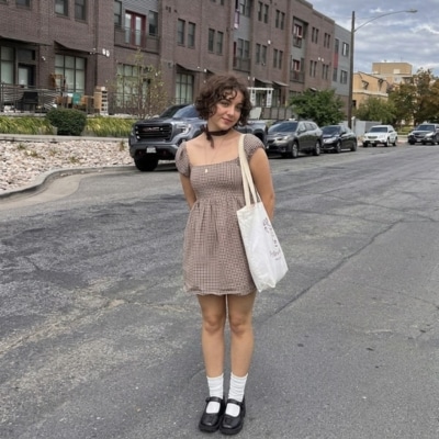 Young woman wearing a brown checkered babydoll dress and Mary Jane shoes standing on a street.