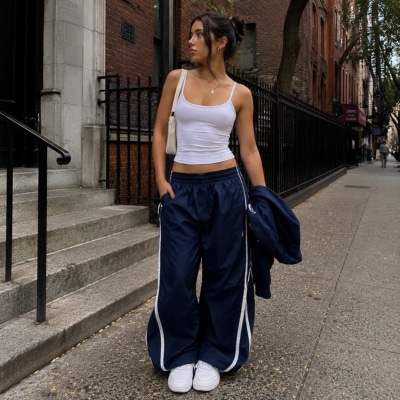 Woman in white crop top and navy track pants standing on city sidewalk with brick buildings and black fence