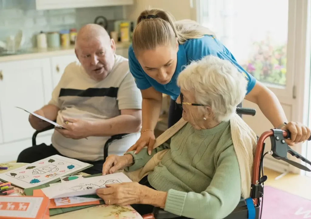 Caregiver assisting two elderly people with coloring pages in a bright room