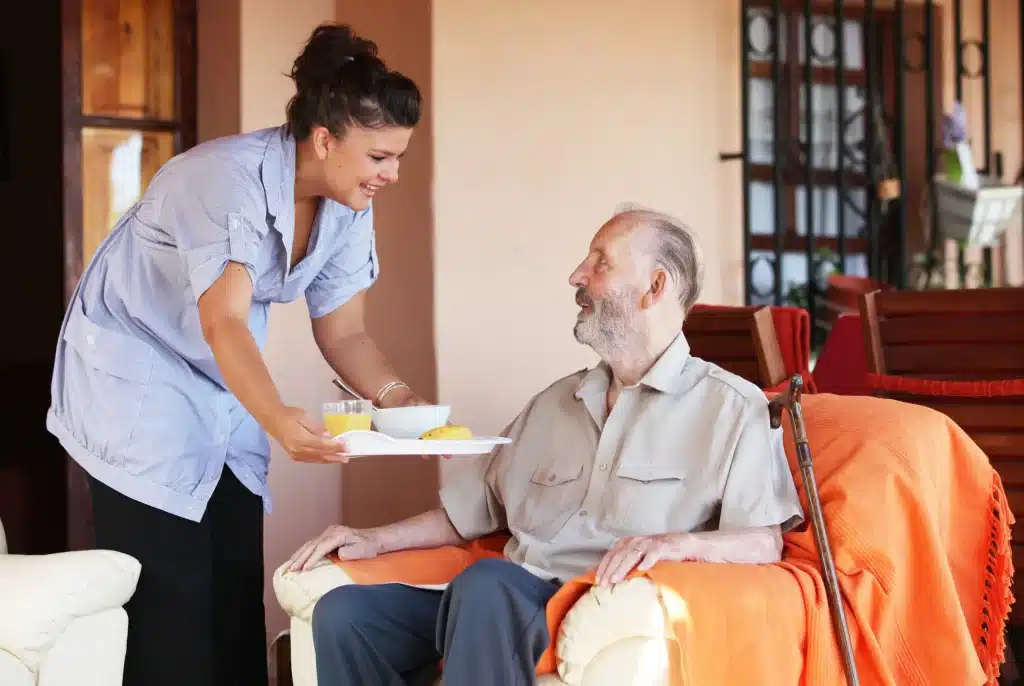 Caregiver serving food to elderly man sitting on chair in warm, outdoor setting