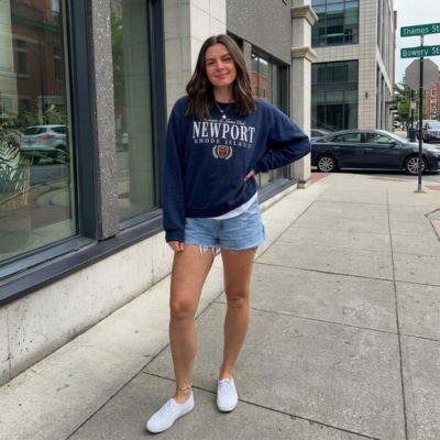 Woman wearing a navy sweatshirt and denim shorts, standing on a city sidewalk with white sneakers.