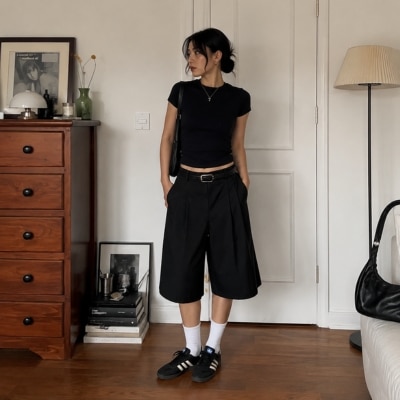 Young woman in black crop top and pleated shorts standing in a cozy, minimalist room with wooden dresser and books.