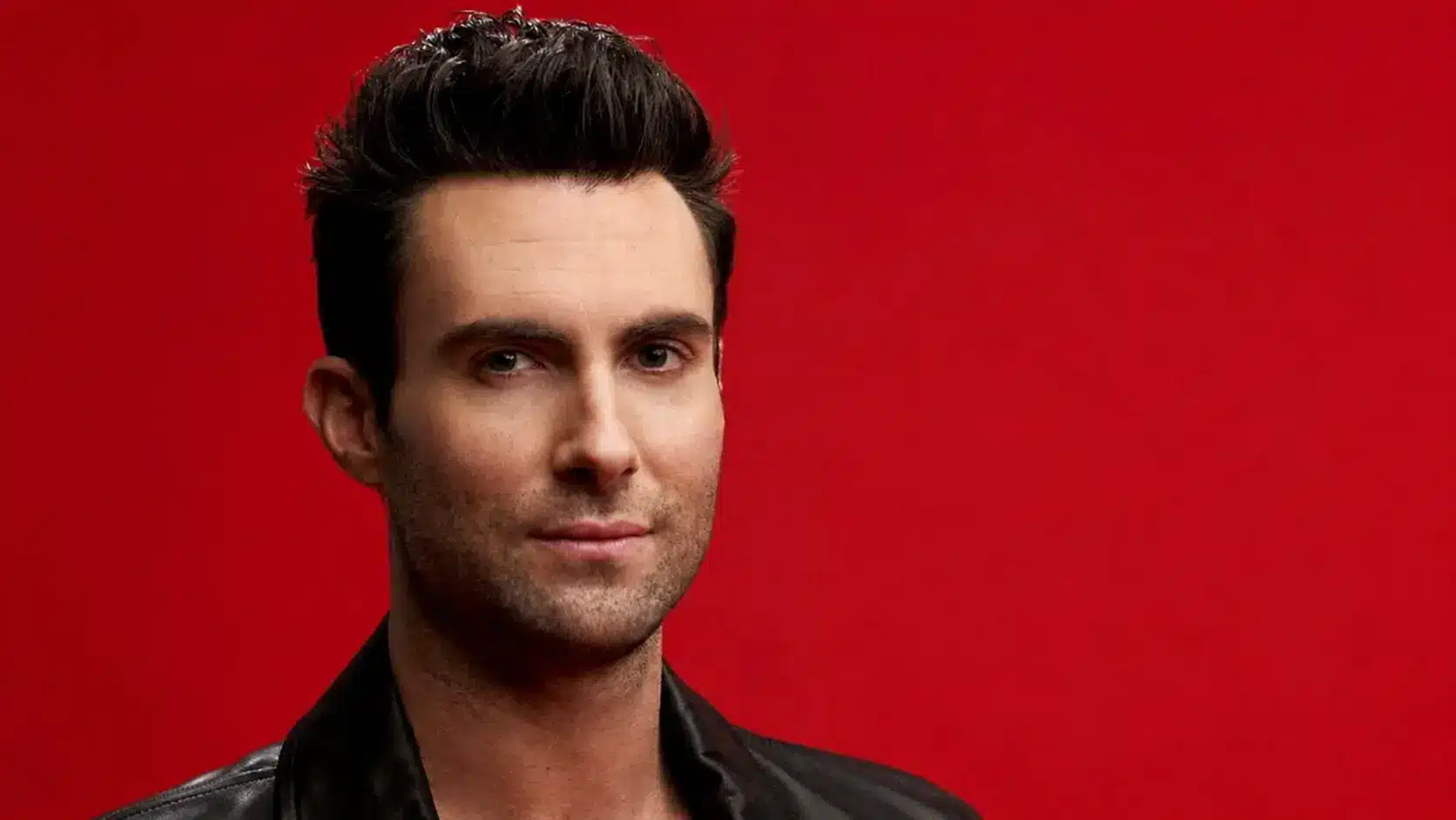Adam levine with styled black hair wearing a black jacket against a bold red background in a portrait shot