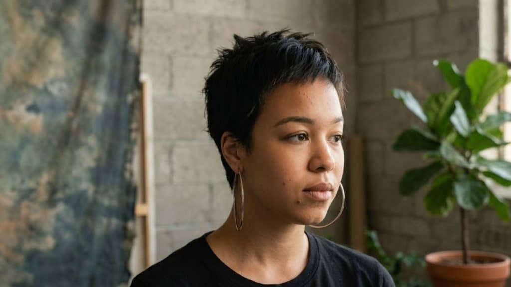 Woman with androgynous haircut having short pixie style, wearing hoop earrings, black tee in indoor plant backdrop.