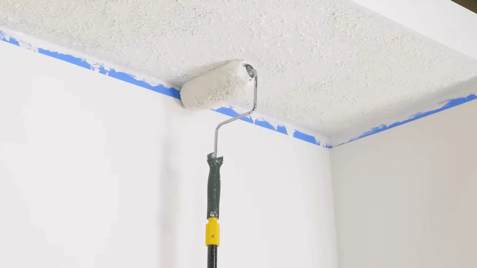 Paint roller applying white paint to a textured popcorn ceiling along taped wall edges.