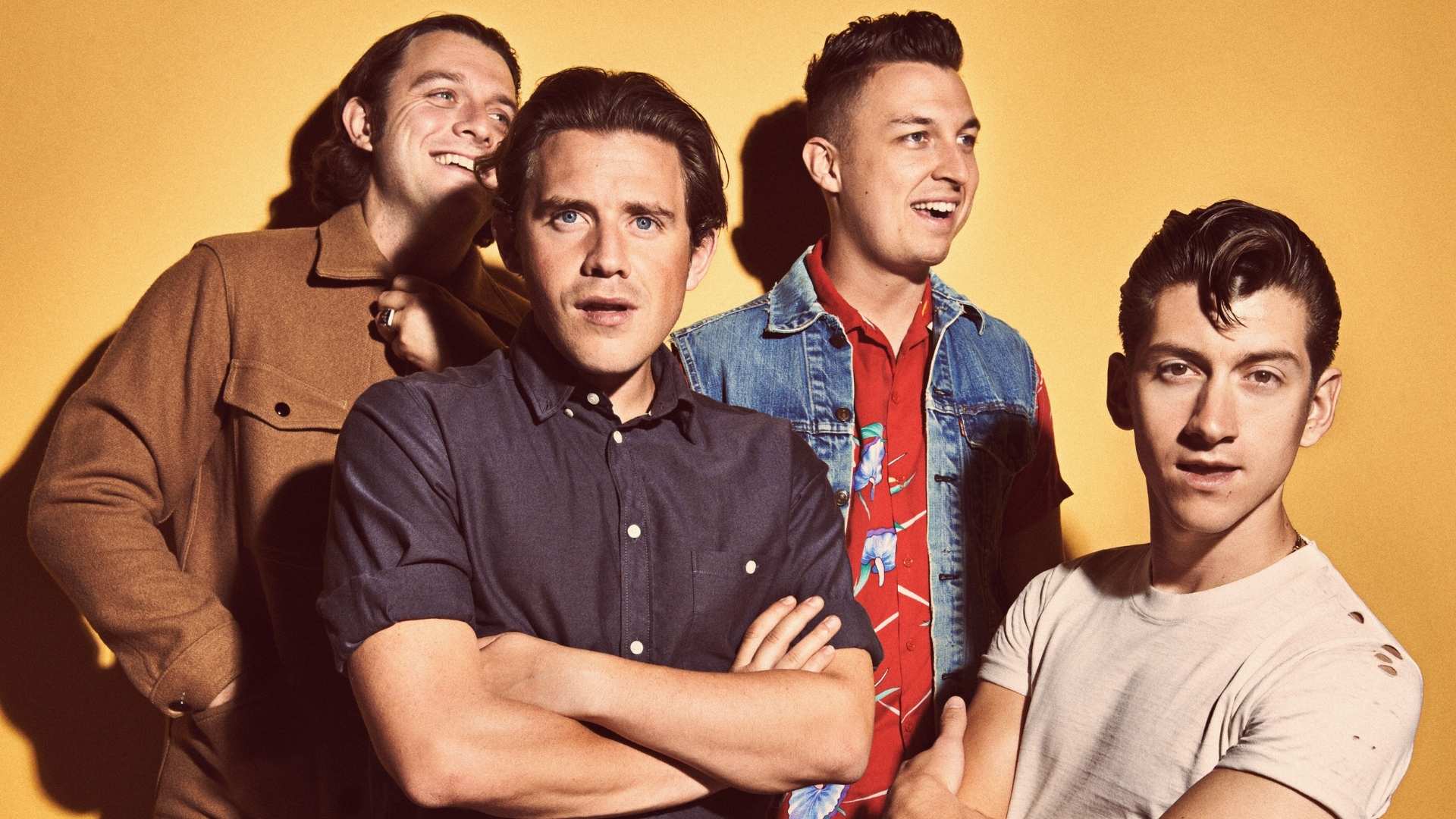 Four indie rock band 'Arctic Monkeys' members posing in studio with arms crossed against warm yellow background, casual confident portrait.