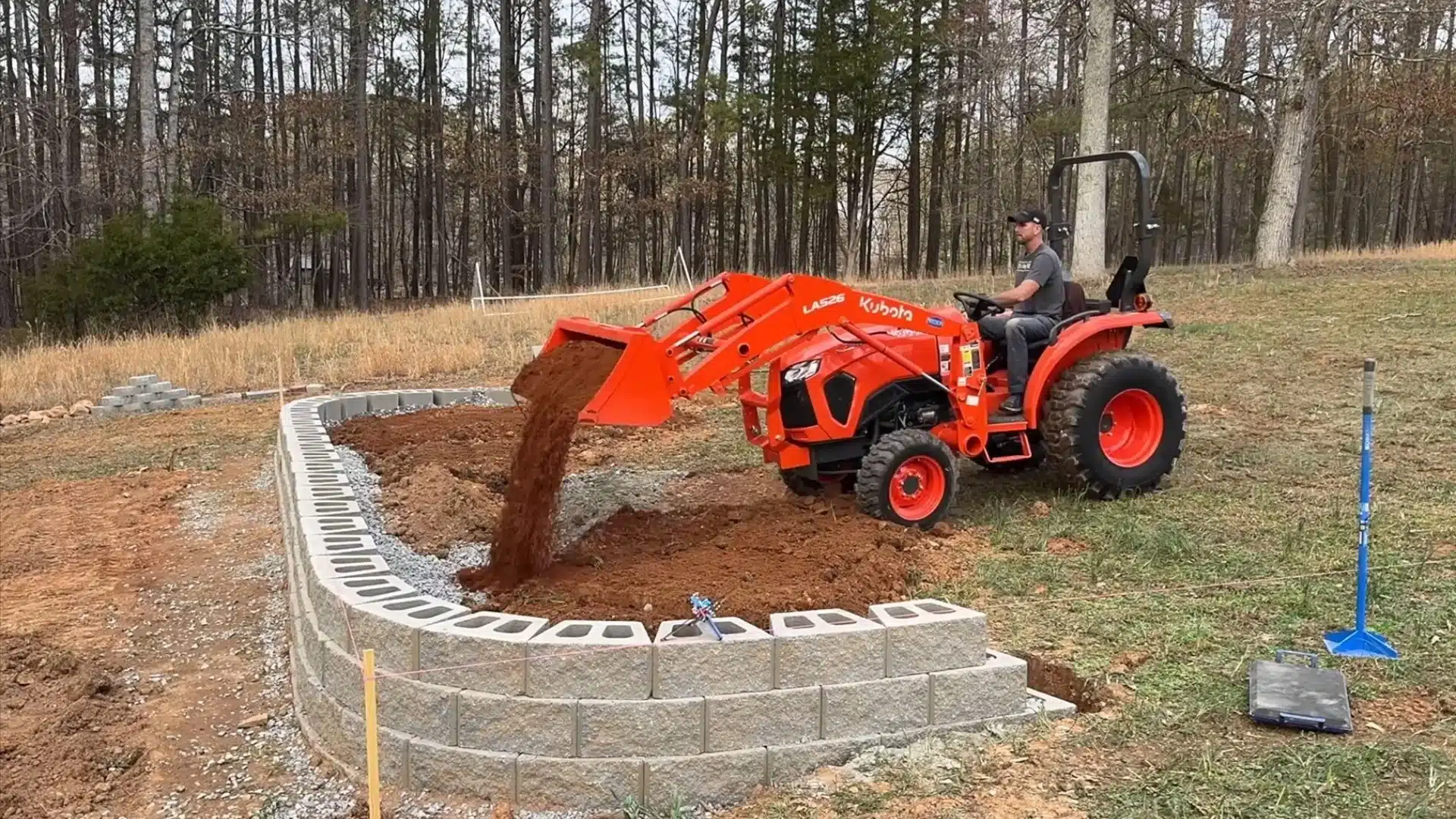 Person using a tractor to move soil while building a curved concrete block retaining wall outdoors.