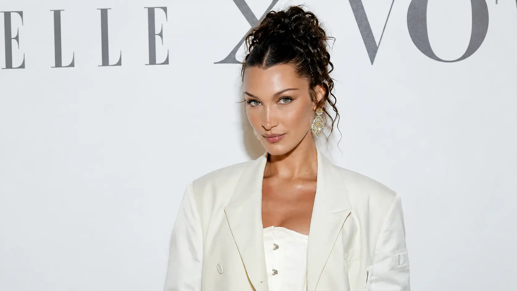 bella hadid wearing a white blazer and updo posing at a fashion event with a minimalist backdrop.