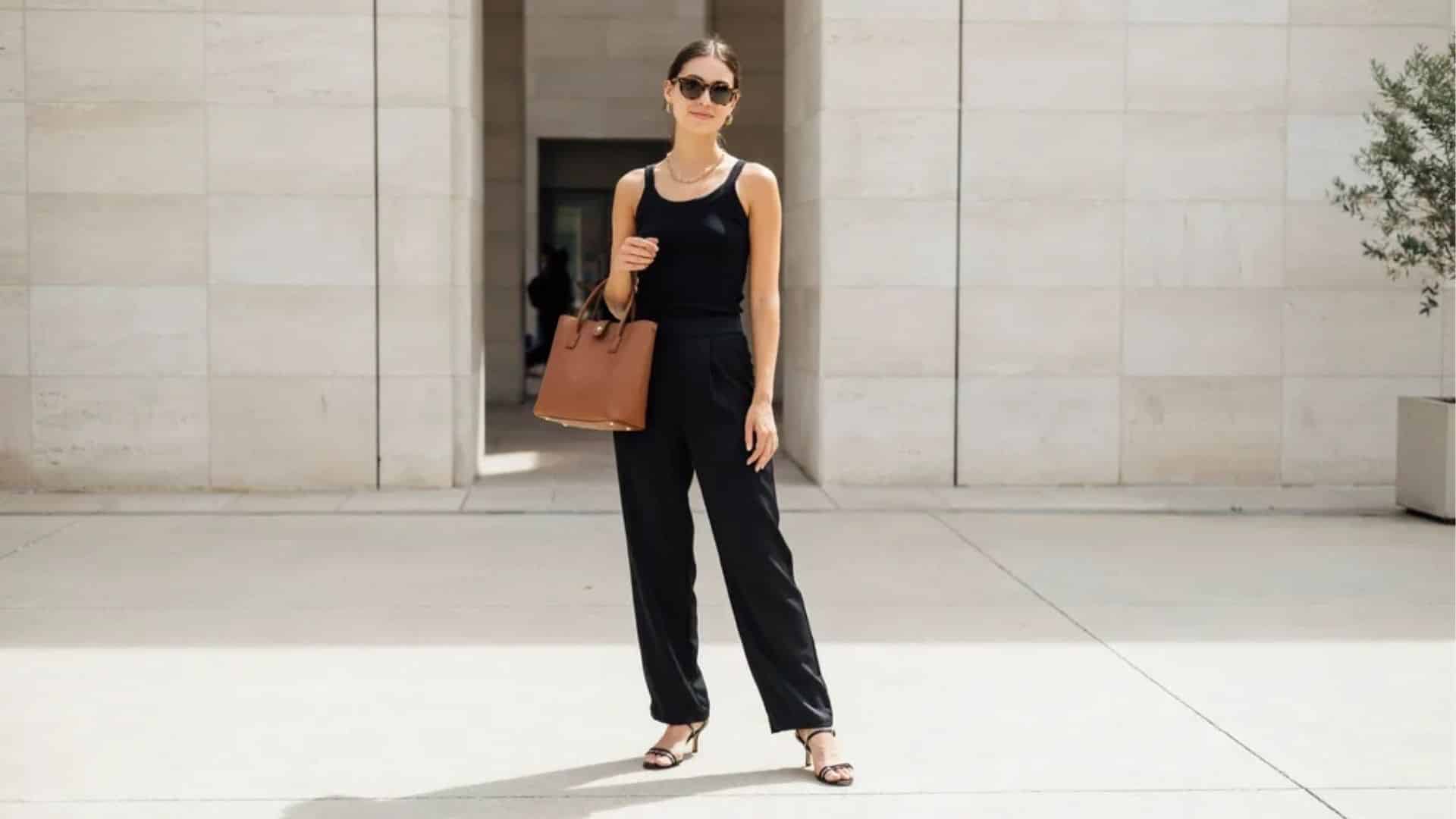 Woman wearing black tank top wide leg trousers and heeled sandals standing on city street with sunglasses and tote bag.
