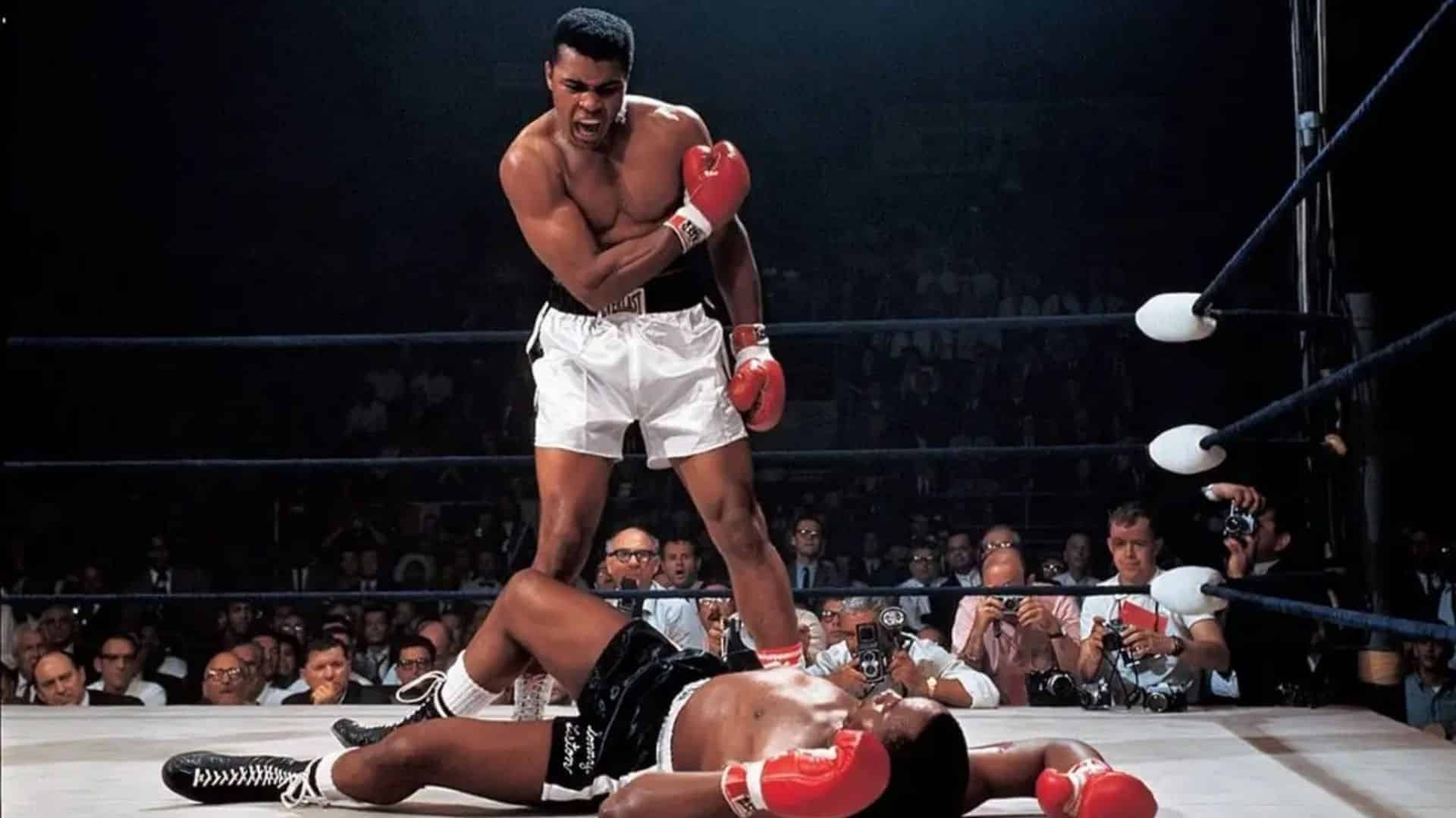 Boxer  muhammad ali in red gloves stands over fallen opponent in boxing ring while crowd and photographers watch