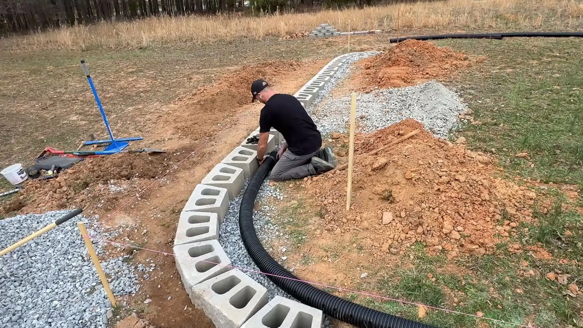 Person installing concrete blocks and drainage pipe along a curved trench during outdoor construction work.